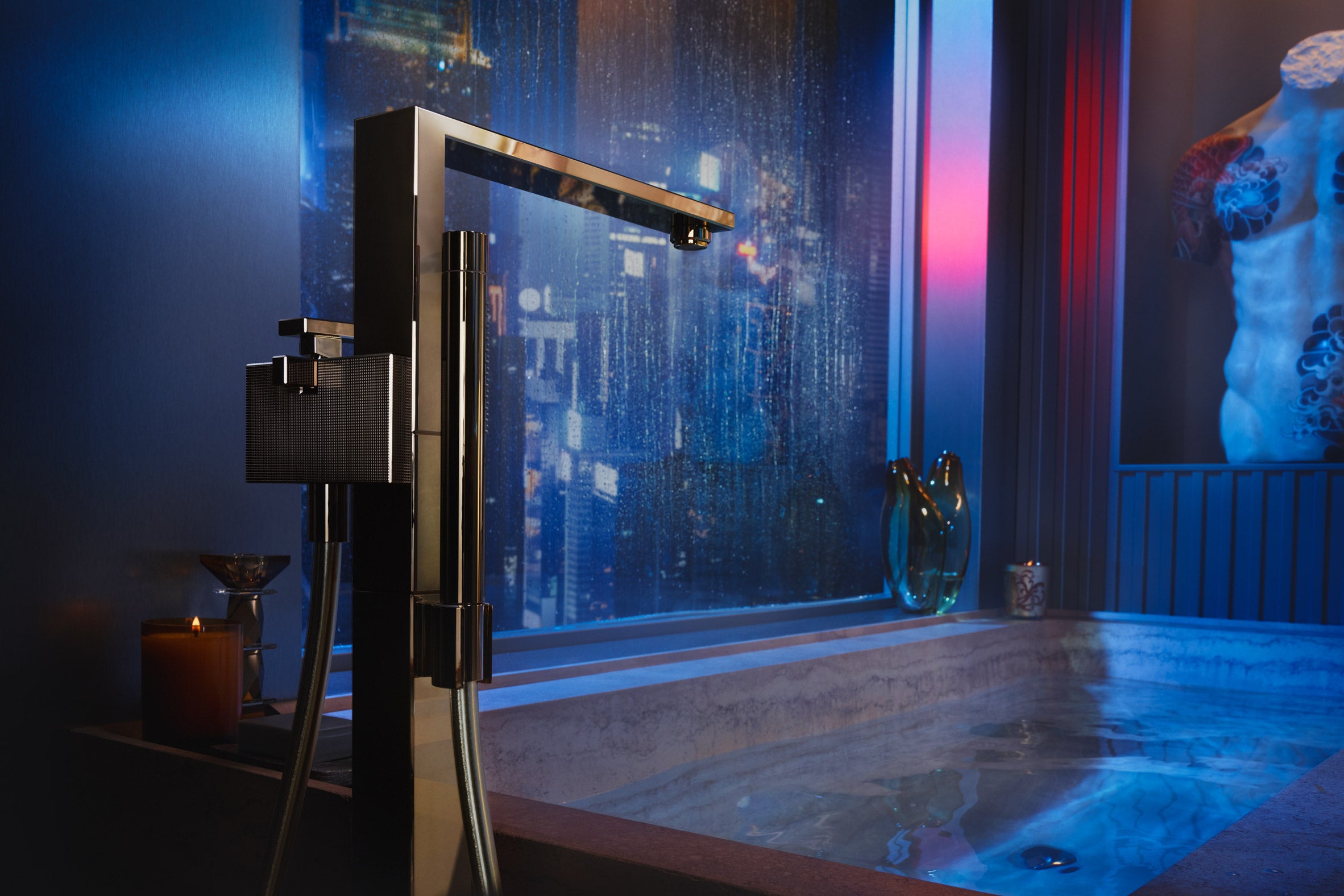 EDGE Gulvstående Badekarsarmatur i Polert Gull fra AXOR, foran marmor badekar. Hele bildet bader i blått neonlys.
