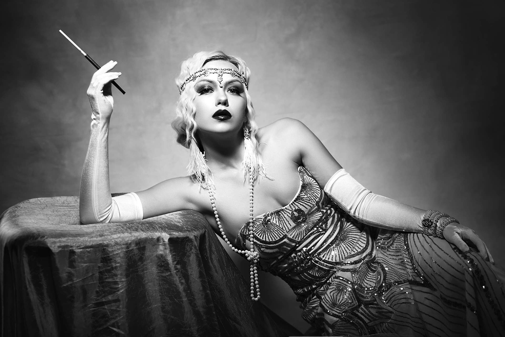 Black and white photo of elegant roaring 20's woman leaning against a table with a cigarette.