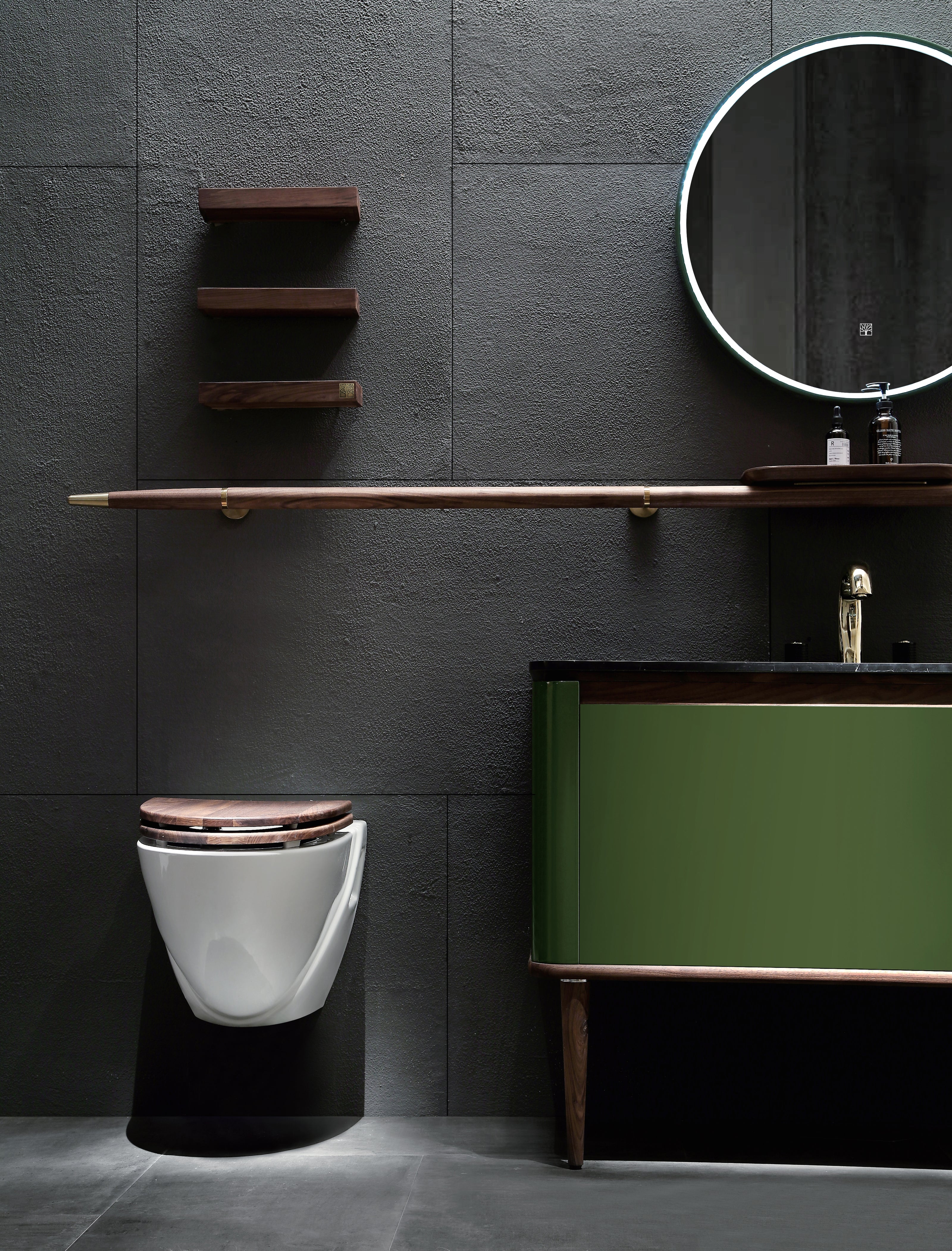 Nærbilde av Eterra baderomsinnredning fra Brandt Bad. Unikt og vakkert møbel med grønn speilblank lakkmaling og detaljer i valnøtt og messing. Servant i sort marmor. Tilbehør er en lang rund håndklestang i valnøtt med en påmontert liten hylle. Rundt speil med integrert LED belysning, og tre vakre vegghyller i valnøtt.