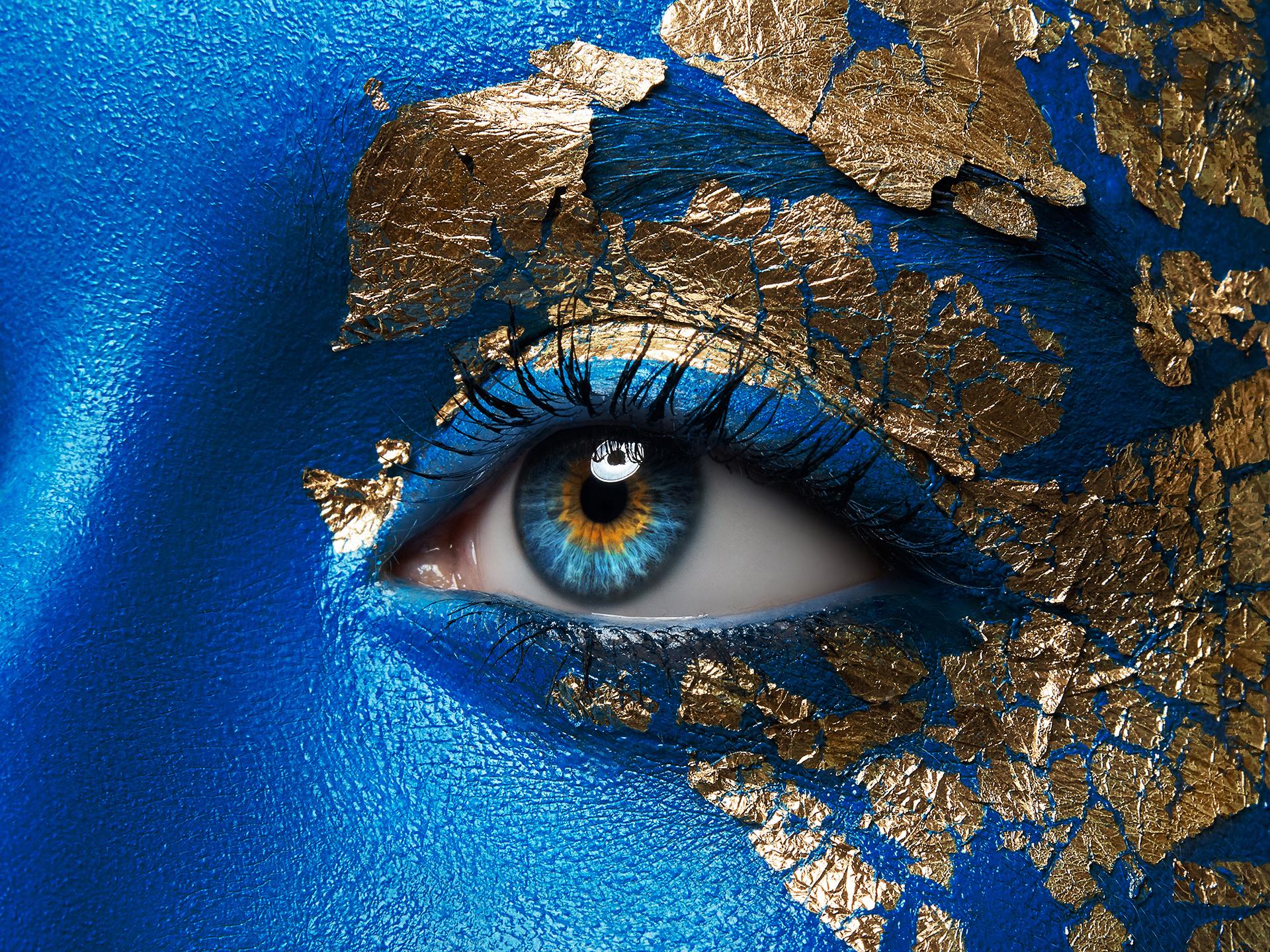 Nærbilde av åpent øye på blåmalt ansikt med gulldetaljer.